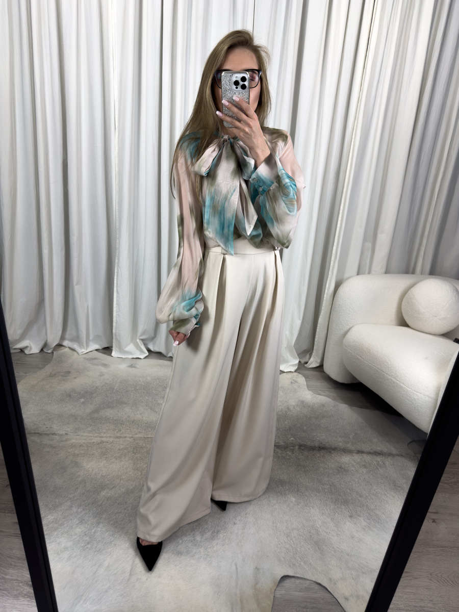 Lśniąca koszula Alice zielona z kokardą przy dekolcie, w odcieniach zieleni i beżu, zestawiona z kremowymi spodniami palazzo – elegancka i świeża stylizacja dla kobiet ceniących klasykę oraz nowoczesny akcent. Modelka na tle białych firan.