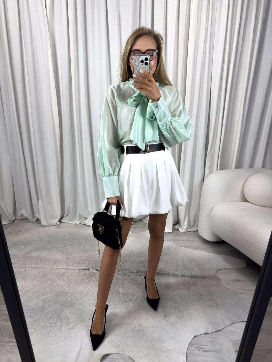 Zdjęcie produktowe sklepu internetowego z odzieżą damską – modelka ubrana w koszule Satine w kolorze pistacjowym/zielonym marki Miho’s, spódnico-spodenki w kolorze białym, trzymająca w ręce małą, czarną torebkę