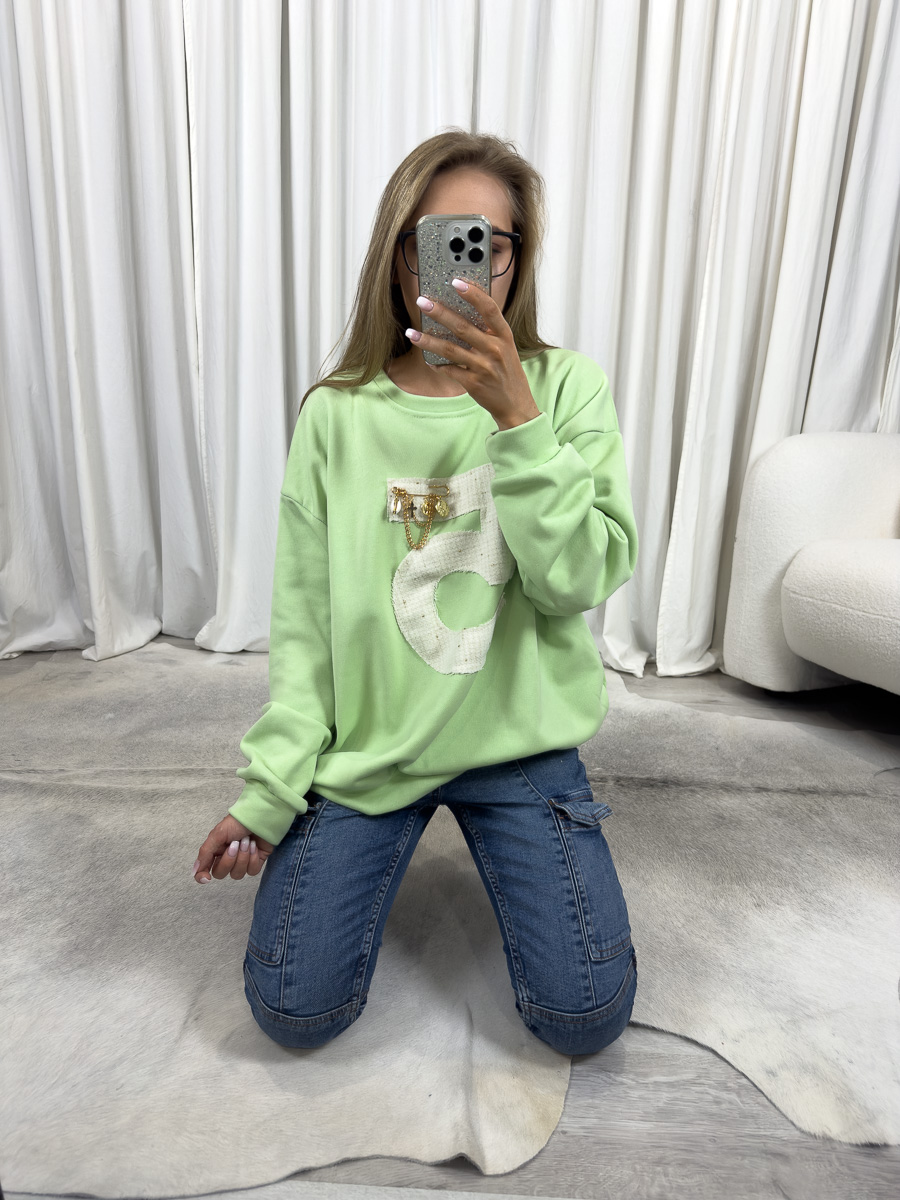 Bluza oversize Five zielona z nadrukiem 5 i ozdobną przypinką, stylizacja z jeansami i białymi sneakersami, zdjęcie produktowe na tle jasnych zasłon.