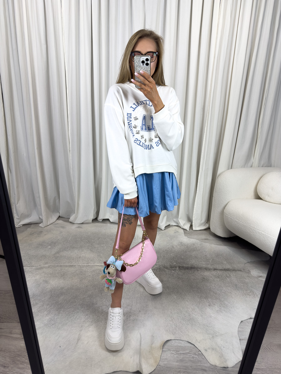 Bluza oversize College biała z nadrukiem college, stylizacja z błękitną spódnicą, białymi sneakersami i różową torebką, zdjęcie produktowe na tle jasnych zasłon.