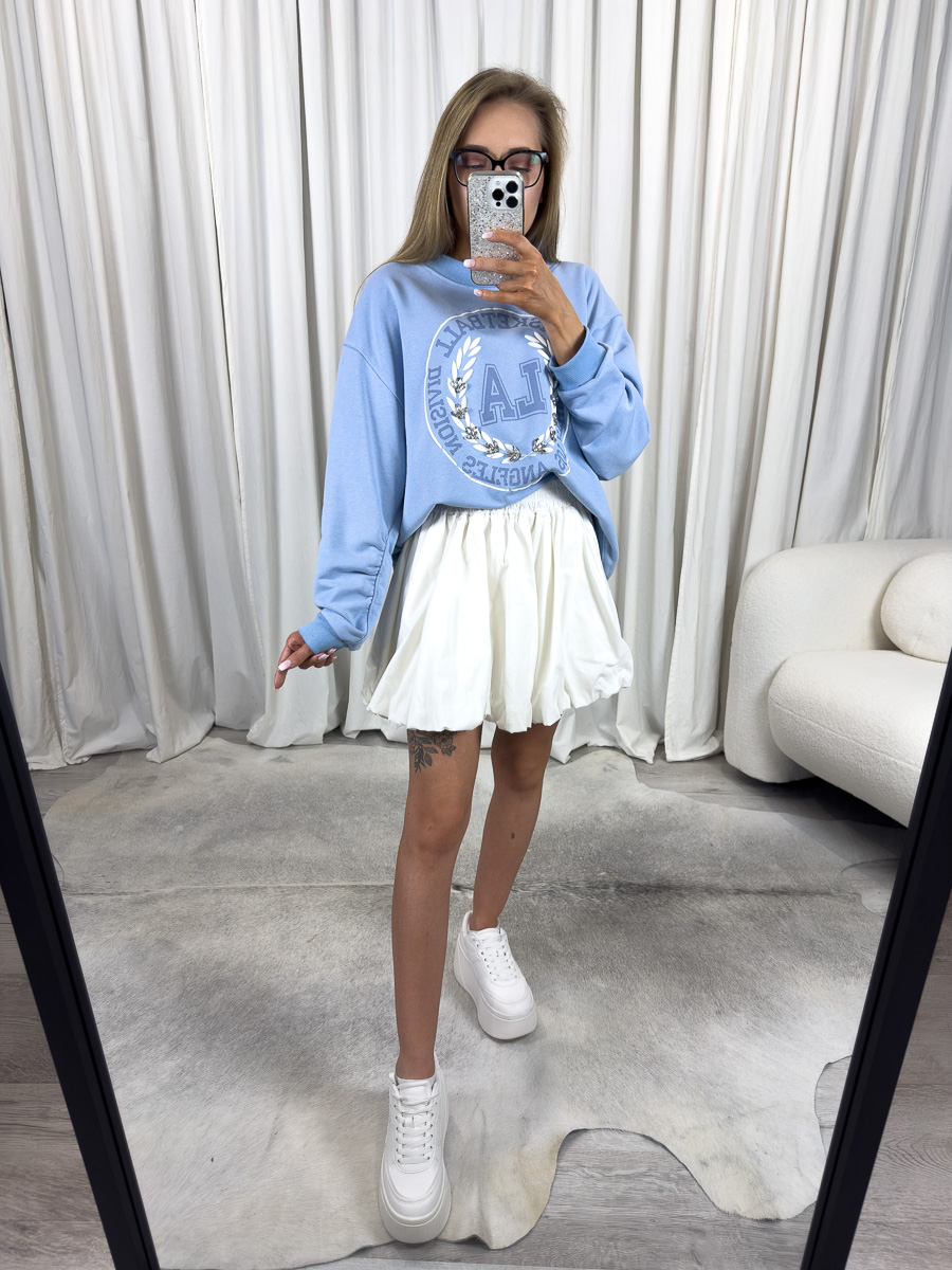 Bluza oversize College błękitna z nadrukiem college, stylizacja z białą spódnicą i białymi sneakersami, zdjęcie produktowe na tle jasnych zasłon