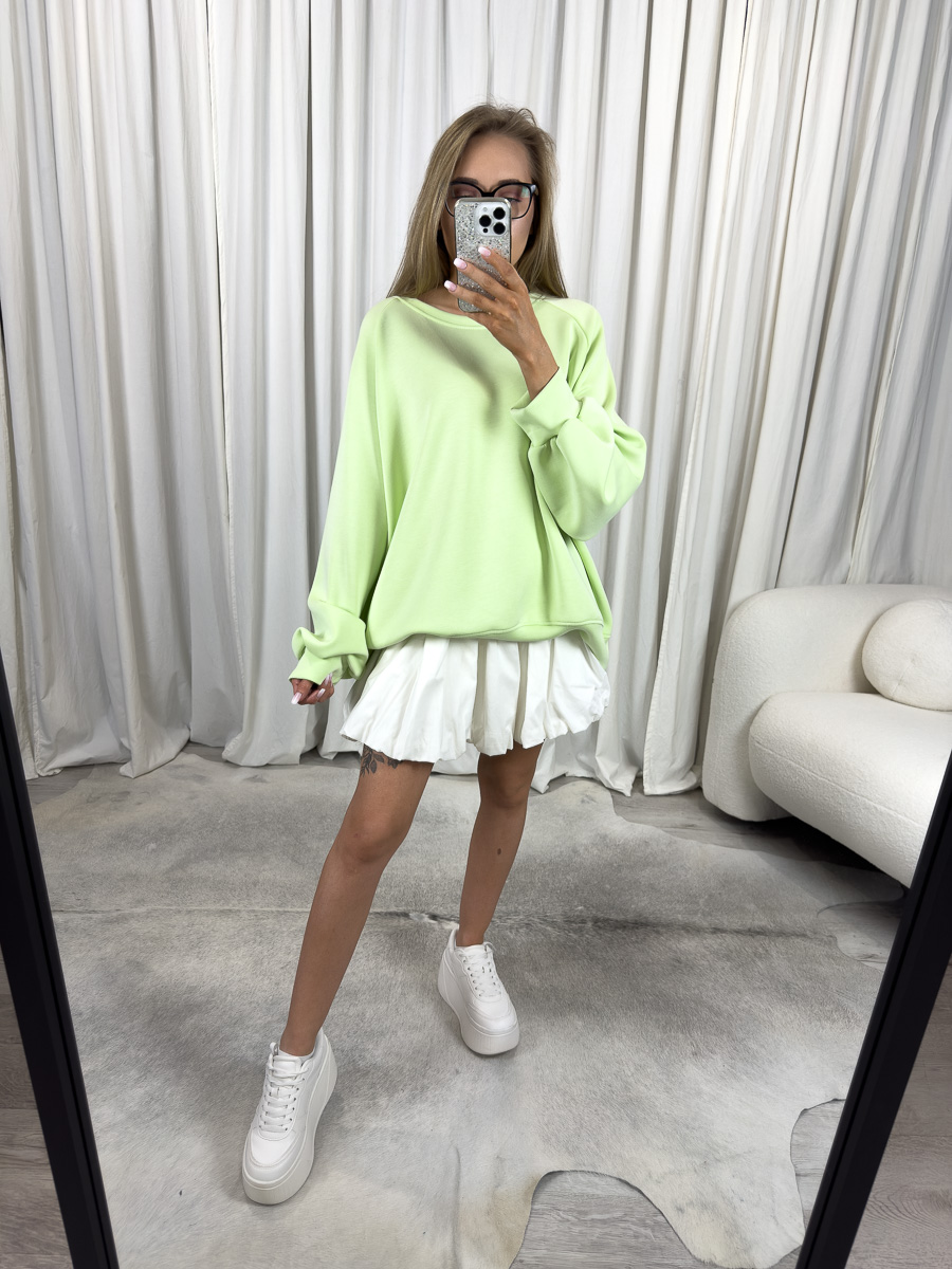 Modelka w jasnozielonej bluzie oversize Casual i plisowanej białej spódnicy, stylizacja streetwear na tle jasnych zasłon.