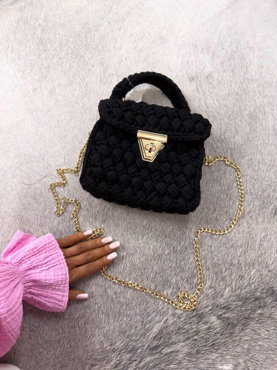 Czarna elegancka torebka Boheme z plecionego sznurka ze złotym łańcuszkiem na tle szarego dywanu ze skóry. Zdjęcie produktowe sklepu internetowego z odzieżą damską.