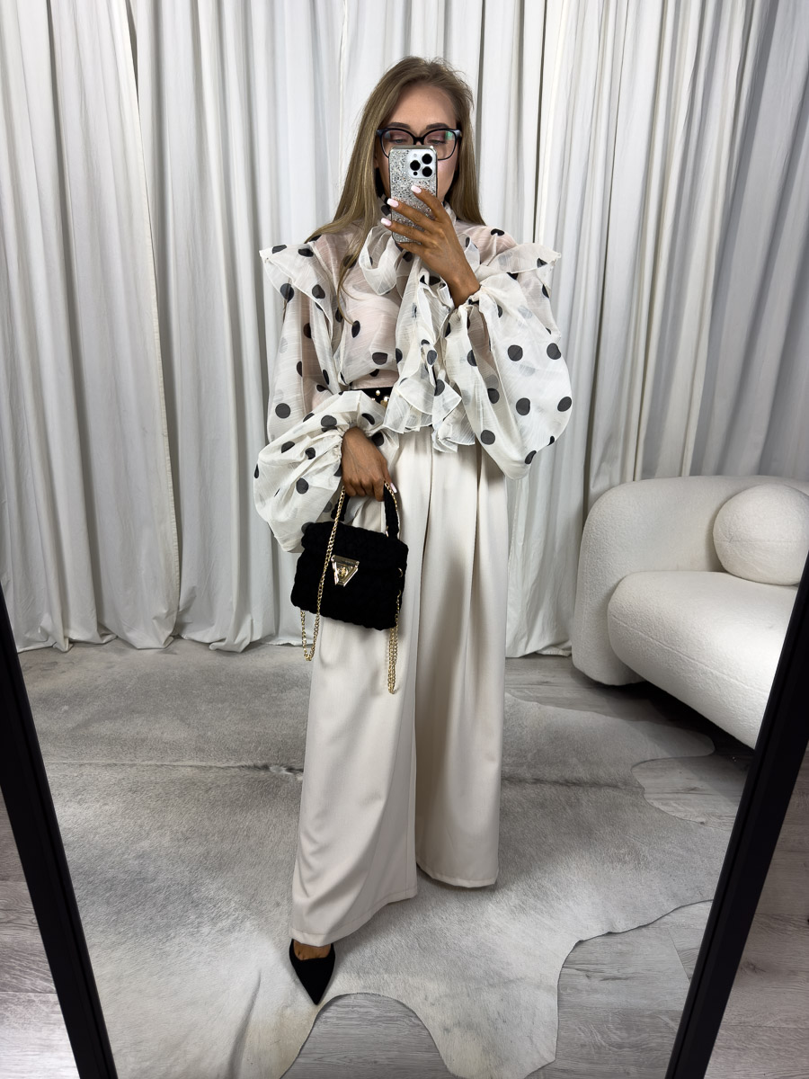 Modelka w eleganckich, kremowych spodniach z szerokimi nogawkami i wysokim stanem, stylizacja z przezroczystą bluzką w grochy, czarną torebką i szpilkami – zdjęcie produktowe do butiku online.