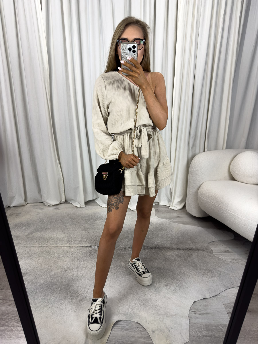 Modelka w muślinowym kombinezonie Girlish z asymetrycznym ramieniem, falbankami i wiązaniem w talii. Zdjęcie produktowe sklepu internetowego z odzieżą damską.