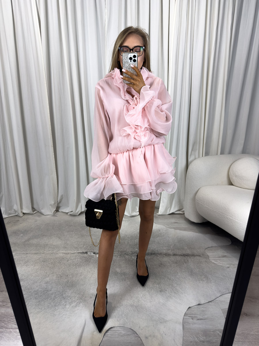 Modelka w sukience mega mini Fleurs z ozdobną aplikacją kwiatową, przezroczystą bluzką i rozkloszowanym dołem, zestawiona z paskiem, szpilkami i czarną torebką – zdjęcie produktowe do sklepu odzieżowego online.